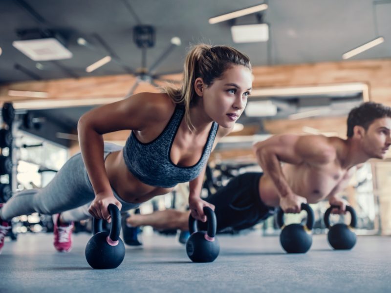 Young couple is working out at gym. Attractive woman and handsome muscular man are training in light modern gym. Doing plank on kettlebell. Push-up on weights.